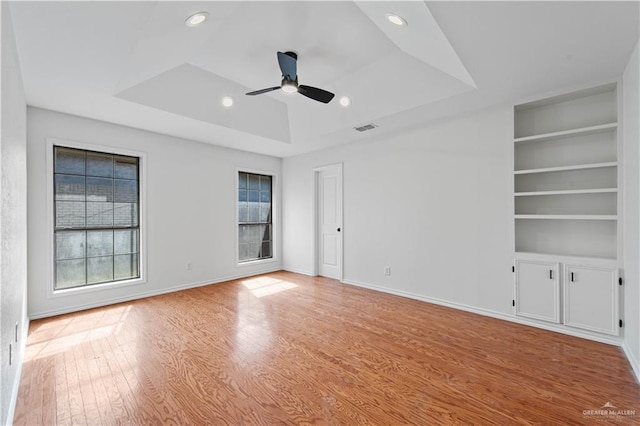 empty room with a raised ceiling, ceiling fan, light hardwood / wood-style floors, and built in shelves