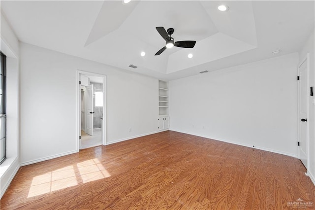 unfurnished room with wood-type flooring, built in features, ceiling fan, and a tray ceiling