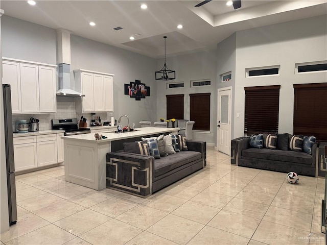 kitchen featuring wall chimney exhaust hood, stainless steel range oven, a towering ceiling, an island with sink, and white cabinets