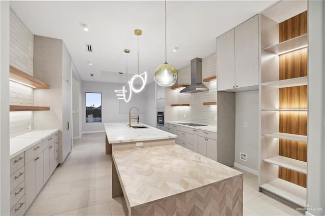 kitchen with an island with sink, black electric stovetop, wall chimney range hood, open shelves, and a sink