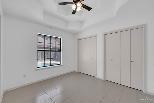 unfurnished bedroom featuring a tray ceiling, light tile patterned floors, multiple closets, ceiling fan, and baseboards