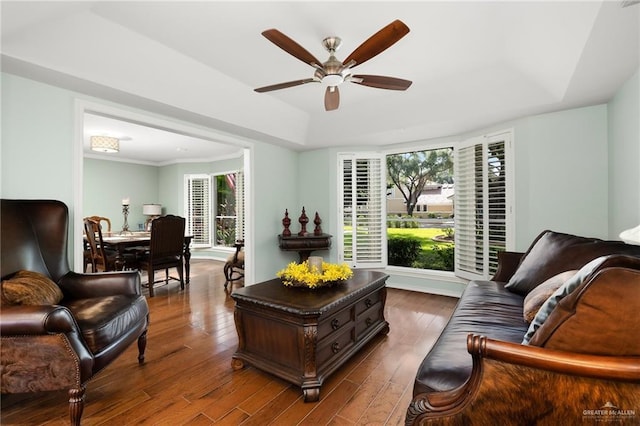 living area with a healthy amount of sunlight, a ceiling fan, a raised ceiling, and dark wood-style flooring