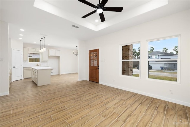 unfurnished living room featuring sink, ceiling fan with notable chandelier, light wood-type flooring, and a tray ceiling