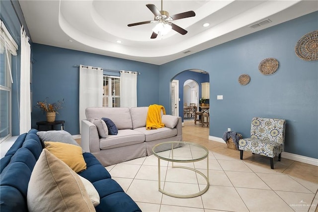 tiled living area featuring arched walkways, a tray ceiling, visible vents, ceiling fan, and baseboards