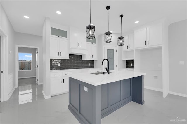 kitchen with tasteful backsplash, sink, decorative light fixtures, white cabinetry, and an island with sink