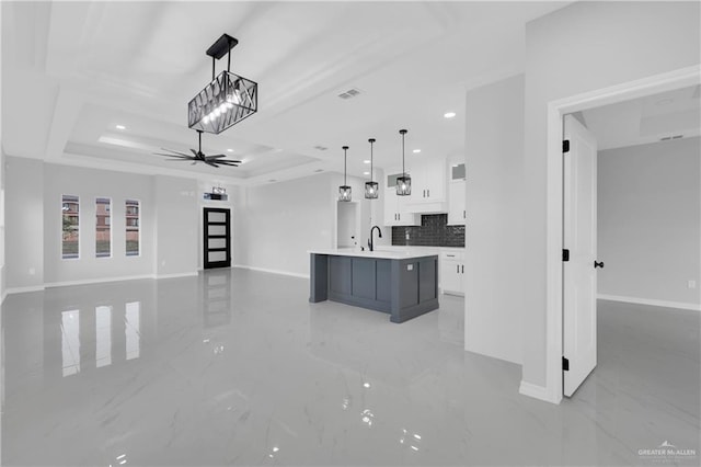 kitchen with white cabinets, a raised ceiling, hanging light fixtures, ceiling fan, and an island with sink