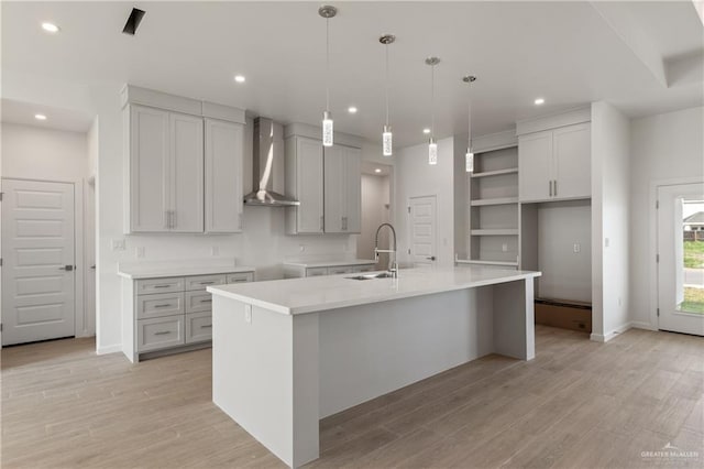 kitchen with recessed lighting, a sink, an island with sink, wall chimney range hood, and light wood-style flooring