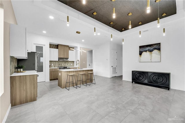 kitchen with backsplash, white cabinets, a kitchen bar, hanging light fixtures, and a center island with sink