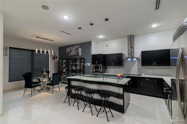 kitchen with decorative light fixtures, tasteful backsplash, an island with sink, stainless steel appliances, and wall chimney range hood