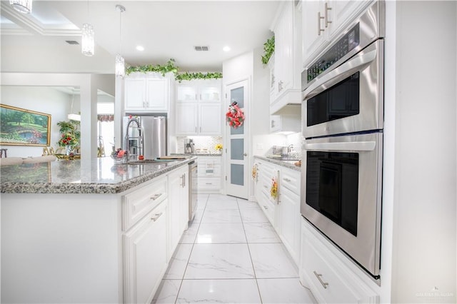 kitchen with appliances with stainless steel finishes, light stone counters, white cabinets, hanging light fixtures, and an island with sink