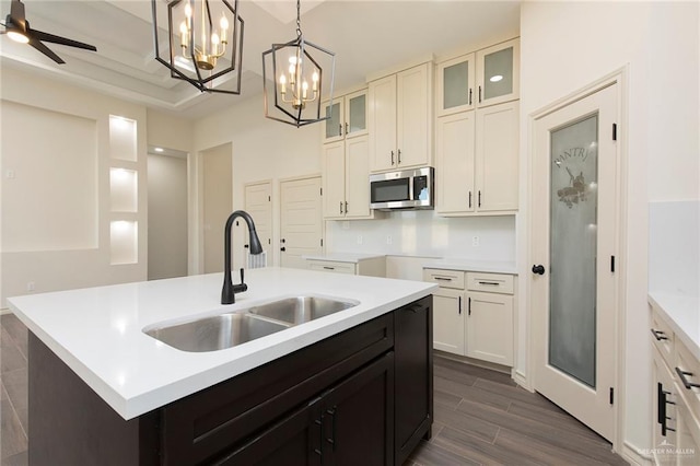 kitchen with stainless steel microwave, glass insert cabinets, an island with sink, hanging light fixtures, and a sink