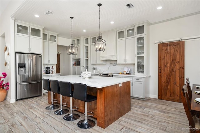 kitchen featuring stainless steel appliances, a barn door, an island with sink, decorative light fixtures, and white cabinets