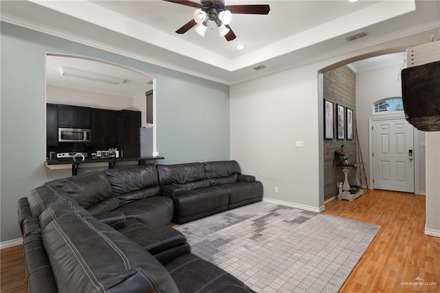 living room with a tray ceiling, light wood-type flooring, visible vents, ceiling fan, and arched walkways