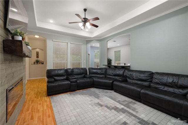 living area featuring a fireplace, ceiling fan, arched walkways, a raised ceiling, and light wood-style floors