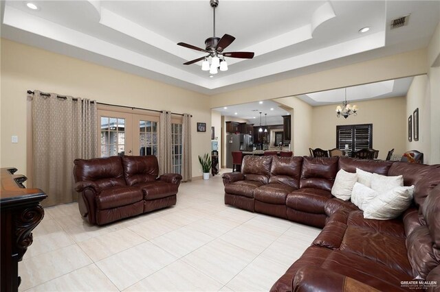 living room with french doors, ceiling fan with notable chandelier, a tray ceiling, and light tile patterned flooring