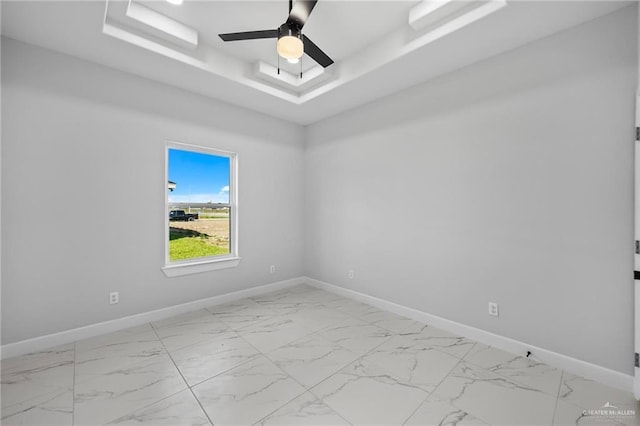 empty room featuring a raised ceiling, light marble finish flooring, ceiling fan, and baseboards