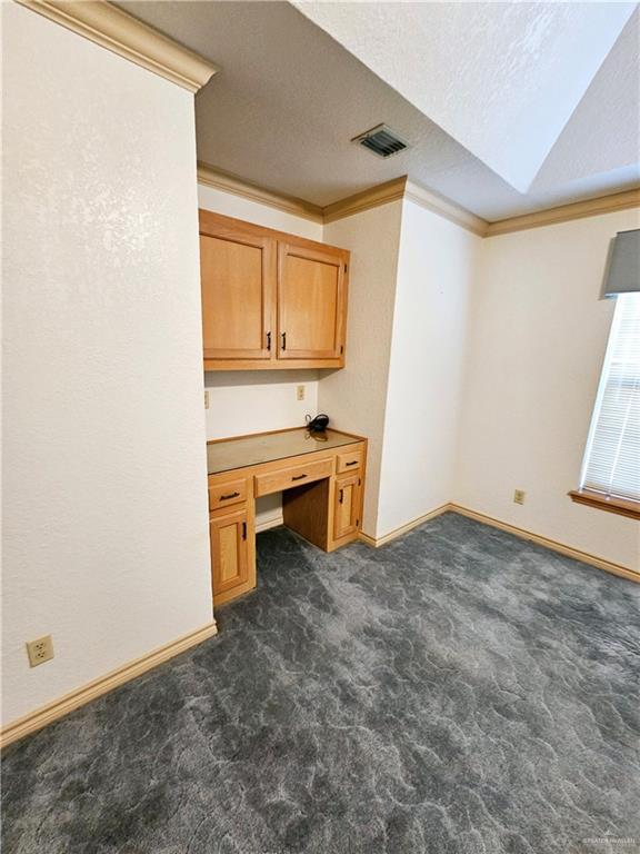 unfurnished office with dark colored carpet, ornamental molding, built in desk, and a textured ceiling