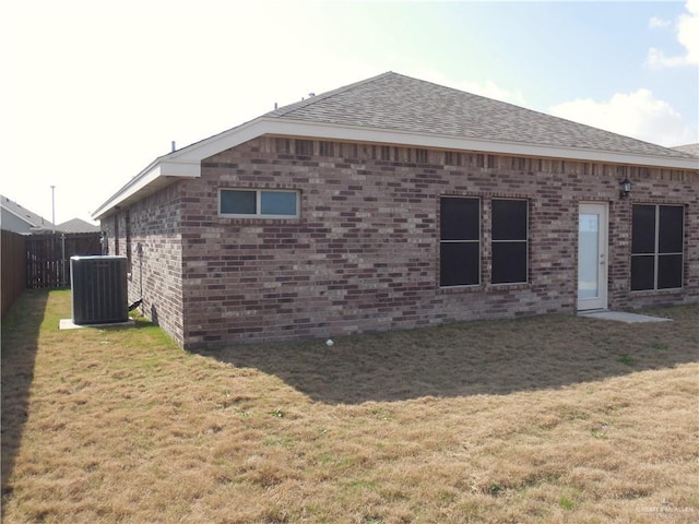 rear view of property with fence, roof with shingles, central air condition unit, a lawn, and brick siding