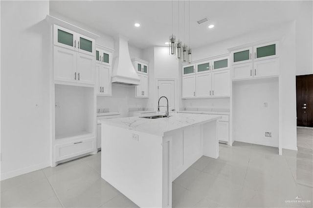 kitchen with a center island with sink, custom range hood, pendant lighting, light stone counters, and sink