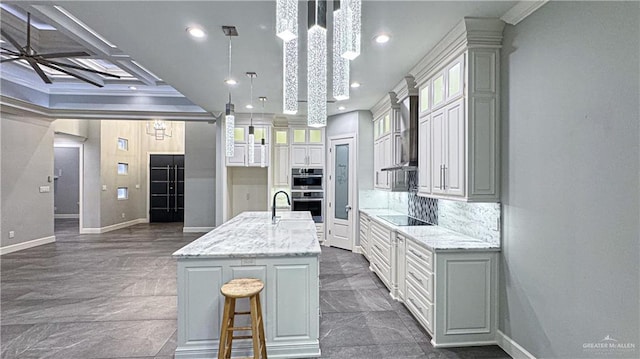 kitchen featuring wall chimney range hood, stainless steel double oven, a kitchen island with sink, and crown molding