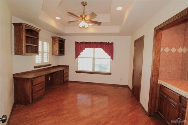 interior space with built in desk, a raised ceiling, ceiling fan, and dark wood-type flooring