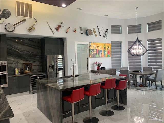 kitchen featuring wine cooler, a large island with sink, stainless steel fridge, dark stone counters, and decorative light fixtures