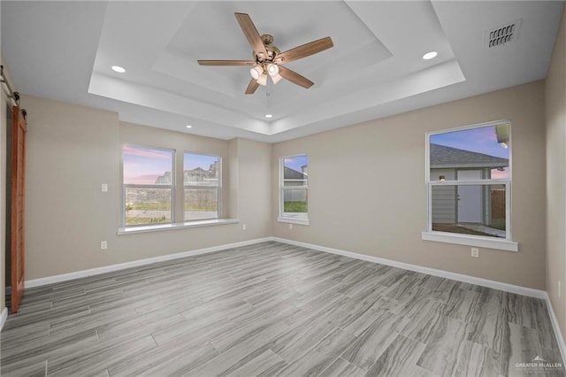 unfurnished room featuring ceiling fan, light hardwood / wood-style floors, a barn door, and a tray ceiling