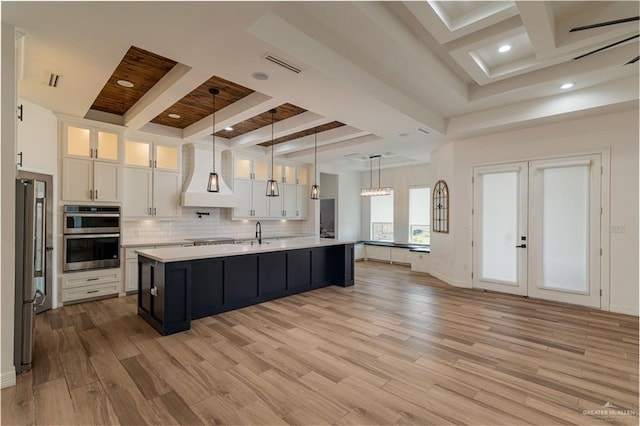 kitchen featuring custom exhaust hood, a kitchen island with sink, hanging light fixtures, appliances with stainless steel finishes, and white cabinetry