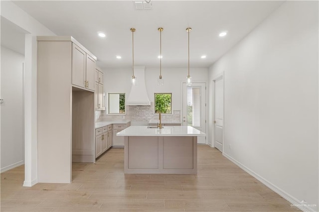 kitchen with decorative light fixtures, tasteful backsplash, an island with sink, custom exhaust hood, and light hardwood / wood-style flooring