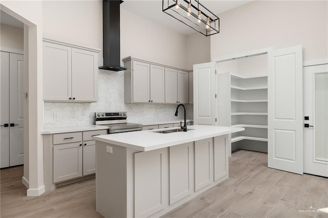 kitchen with wall chimney range hood, sink, electric stove, a center island with sink, and hanging light fixtures