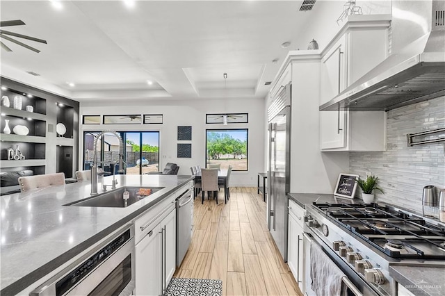 kitchen featuring wall chimney exhaust hood, built in appliances, a sink, ceiling fan, and a tray ceiling