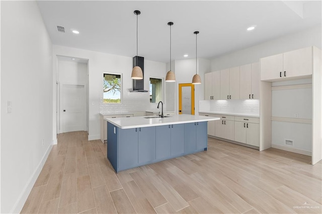 kitchen featuring visible vents, an island with sink, a sink, wall chimney range hood, and light countertops