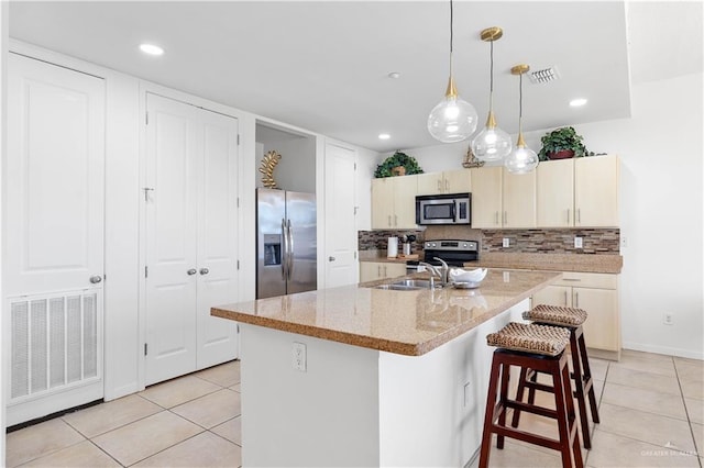 kitchen featuring light stone counters, cream cabinets, an island with sink, decorative backsplash, and appliances with stainless steel finishes