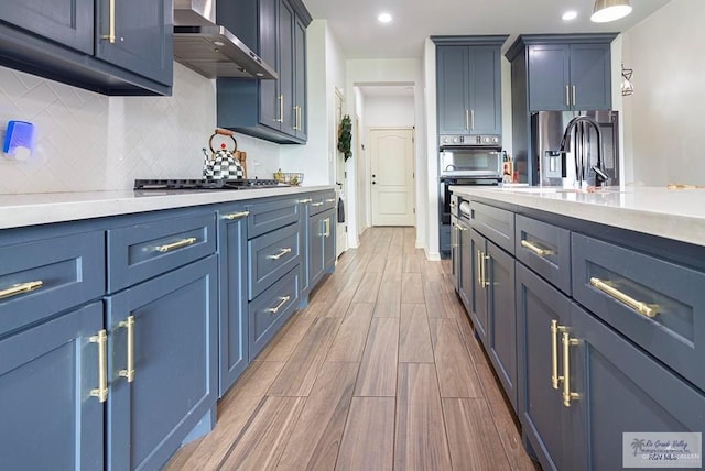 9753 Lake Arthur Ct, Los Fresnos, TX 78566 - kitchen with wall chimney exhaust hood, appliances with stainless steel finishes, blue cabinetry, wood finished floors, and backsplash