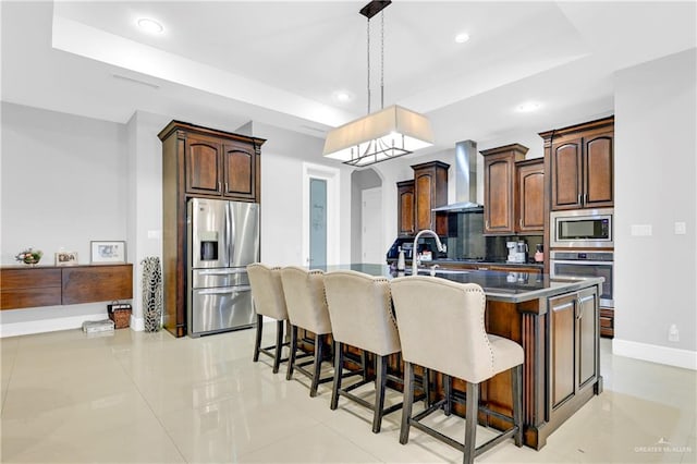 kitchen featuring stainless steel appliances, wall chimney range hood, an island with sink, decorative light fixtures, and a breakfast bar