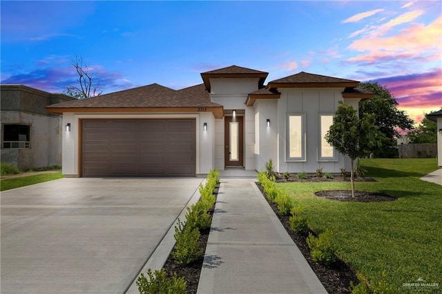 prairie-style home featuring an attached garage, stucco siding, roof with shingles, a yard, and driveway