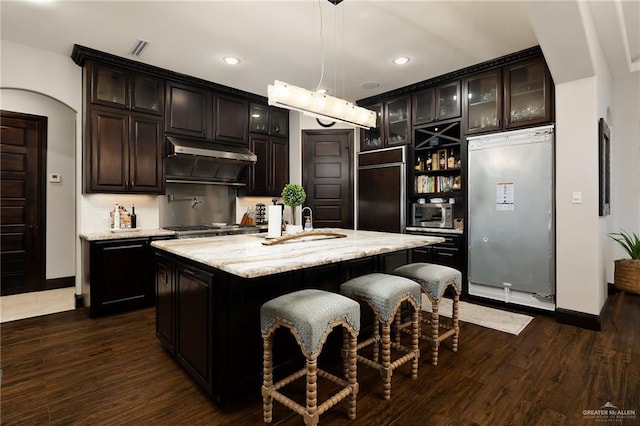 kitchen with dark brown cabinetry, hanging light fixtures, dark hardwood / wood-style floors, built in appliances, and an island with sink