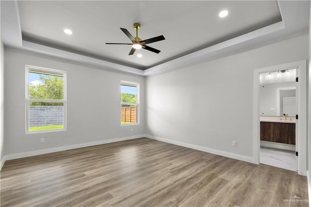 unfurnished bedroom with a raised ceiling, light wood-type flooring, baseboards, connected bathroom, and recessed lighting