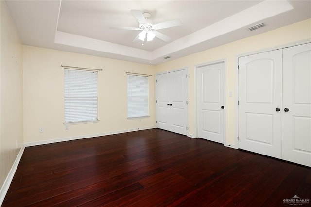 unfurnished bedroom with a raised ceiling, two closets, ceiling fan, and dark hardwood / wood-style floors