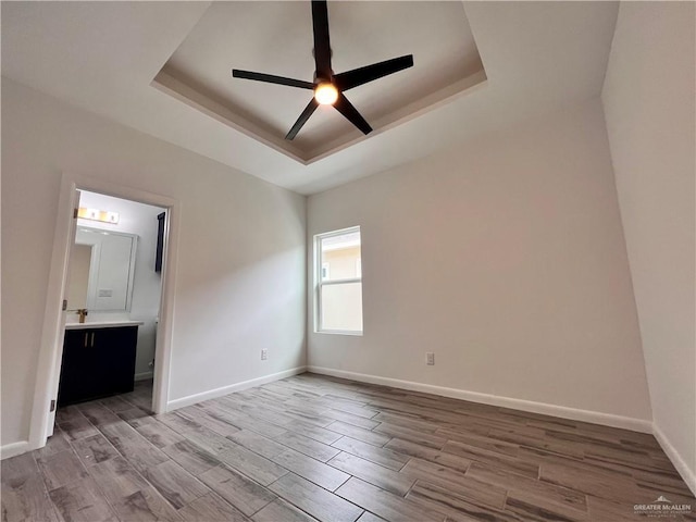 unfurnished bedroom featuring a ceiling fan, ensuite bathroom, light wood-type flooring, a raised ceiling, and baseboards