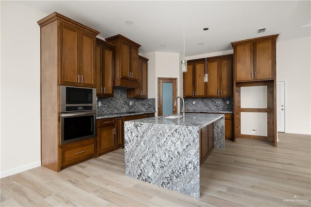 kitchen with stainless steel appliances, tasteful backsplash, light stone counters, a center island with sink, and light wood-type flooring