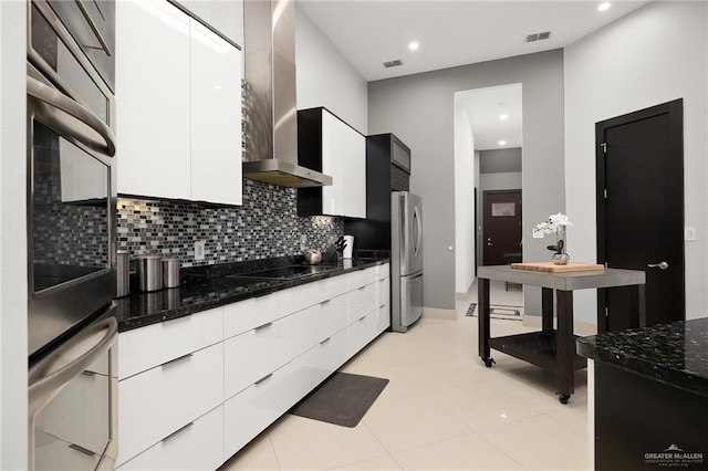 kitchen featuring backsplash, wall chimney exhaust hood, stainless steel appliances, dark stone countertops, and white cabinets