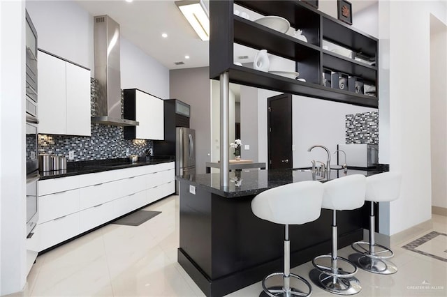 kitchen featuring white cabinetry, wall chimney range hood, decorative backsplash, a breakfast bar, and light tile patterned flooring