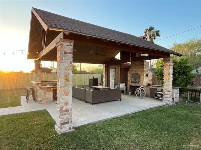 patio terrace at dusk with a lawn, an outdoor living space with a fireplace, fence, a gazebo, and ceiling fan