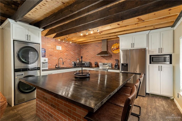 kitchen featuring stacked washer / dryer, dark countertops, wall chimney exhaust hood, appliances with stainless steel finishes, and beam ceiling