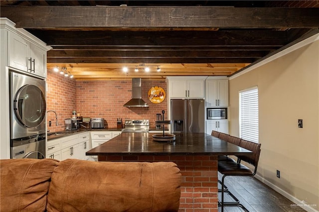 kitchen featuring stacked washer / drying machine, appliances with stainless steel finishes, wall chimney range hood, beam ceiling, and dark countertops