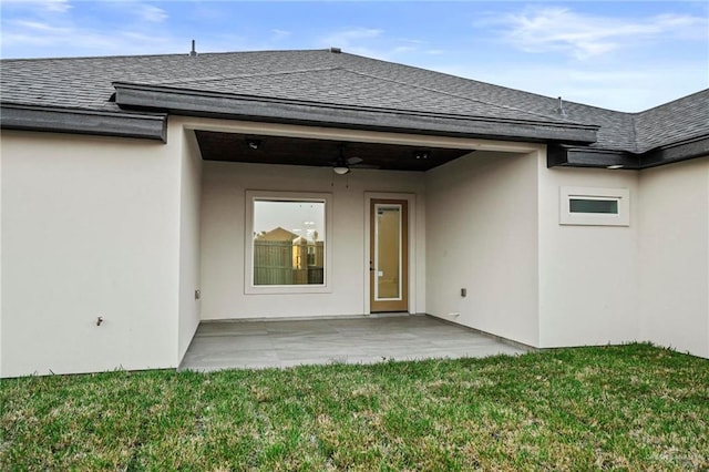view of exterior entry featuring stucco siding, a yard, roof with shingles, and ceiling fan