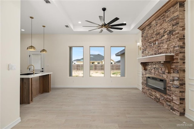 unfurnished living room with a stone fireplace, sink, light hardwood / wood-style flooring, ceiling fan, and a tray ceiling