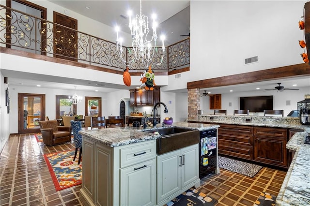 kitchen with a center island with sink, ceiling fan with notable chandelier, sink, wine cooler, and a towering ceiling