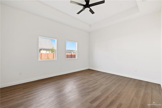 empty room featuring dark wood-style floors, a tray ceiling, a ceiling fan, and baseboards